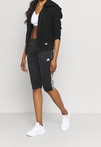 Žena v černé bundě Adidas, černých Adidas kraťasech po kolena se bílými pruhy, bílém sportovním podprsenkovém topu a bílých teniskách, stojící na šedé podlaze.