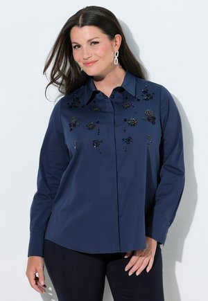 Frau mit langen dunklen Haaren trägt eine marineblaue Bluse mit schwarzen Perlenverzierungen, kombiniert mit schwarzen Hosen und geometrischen Ohrringen, leicht lächelnd.