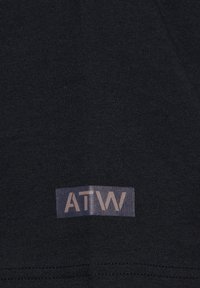 Tissu noir avec une petite étiquette rectangulaire comportant les lettres "ATW" en marron clair sur un fond sombre, près du bord inférieur.