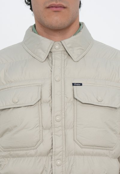 Beige, quilted pufferjakke med spids krave, to brystlommer og knaplukning. Har et "Polo"-logo på venstre lomme.