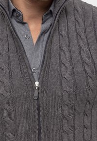 Grijze gebreide vest met kabelpatronen, voorzien van een rits en een grijze shirt met kraag eronder. Gelaagd materiaal met een geribde afwerking.