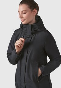 Črna kapuca jakna iz gladkega materiala s sprednjim zadrgo, nastavljivimi vrvicami in dvema stranskima žepoma. Ima visok ovratnik.