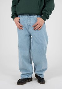 Helerohelised teksapüksid laia lõikega, millel on klassikaline viie taskuga disain ja õmmeldud detailid, kantud mustade kingadega.