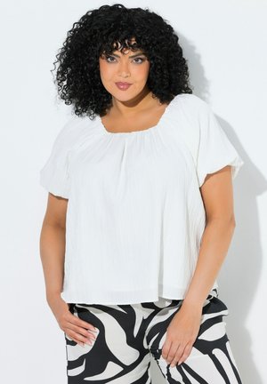 Kobieta o kręconych czarnych włosach, ubrana w luźną białą bluzkę z krótkim rękawem oraz czarno-białe spodnie w wzory, stojąca na jednolitym tle.