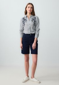Gestreiftes Hemd mit floralem Stickerei auf der Brust, kombiniert mit dunkelblauen Shorts und weißen Sneakers, vor einem schlichten Hintergrund stehend.