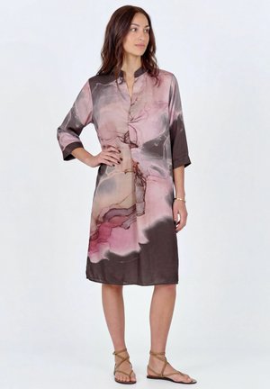 Vrouw draagt een jurk tot op de knie met een abstract patroon in roze, beige en bruin in aquarelstijl, driekwart mouwen, en staat met een hand op haar heup.