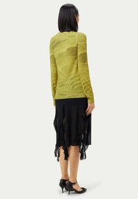 Långärmad, olivgrön tröja med en vågig textur, kombinerad med en svart, volangkjol. Klädesplaggen kompletteras av svarta, spetsiga klackskor.