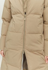 Beige padded coat with a front zipper, featuring a smooth fabric texture and quilted design, displaying minimalistic accents and a relaxed fit.
