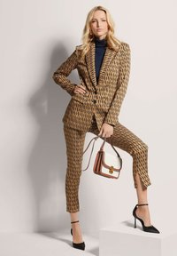 Op maat gemaakt bruin pak met zwart patroon, aansluitend ontwerp en cropped broek; gecombineerd met een zwarte coltrui en zwarte hakken, terwijl een chique handtas wordt vastgehouden.