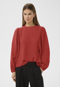 Femme portant un chemisier rouge ample à manches longues avec des épaules bouffantes et un pantalon noir taille haute, debout avec une main dans la poche.