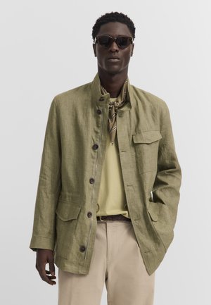 Man wearing sunglasses, olive green buttoned jacket with pockets, beige pants, and a striped neck scarf against a plain white background.