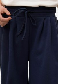 Pantaloni blu navy con tessuto strutturato, dotati di vita con cordoncino e dettagli plissettati sul davanti. Inquadratura ravvicinata dell'area della vita.