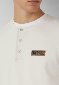Crème kleurige ribgebreide longsleeve shirt met drie metalen knopen en een bruine leren patch met tekst op de borst.
