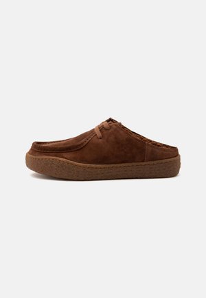 Brun ruskinds slip-on sko med en tyk, tekstureret sål, mokkasin-inspireret syning og snøredetalje, vist i profil på en ensfarvet baggrund.