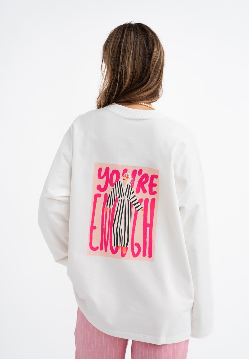 Witte sweatshirt met lange mouwen, voorzien van een felroze grafische print met de tekst "JIJ BENT GENOEG" en een figuur in zwart-witte strepen op de achterkant.