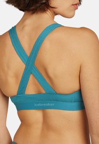 Teal justerbar sportbehå med korsade bärande remmar och ett bekvämt band, med "icebreaker"-logotypen på bandet. Mjuk textur.