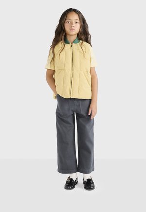 Geel gewatteerd vest met een rits aan de voorkant, korte mouwen, gecombineerd met grijze wijde broek en zwarte instapschoenen met witte sokken.