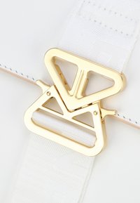 Borsa a tracolla bianca in pelle con superficie testurizzata, dotata di una grande fibbia dorata con un design geometrico e una tracolla in tessuto bianco.