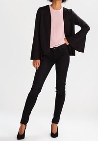 Svart jacka med trumpetärmar över en ljusrosa topp, kombinerad med svarta smala jeans och svarta högklackade skor. Slät tyg med en figurnära design.