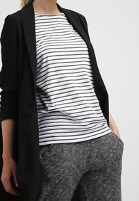 Schwarzer Blazer über einem grau-weißen gestreiften T-Shirt mit rundem Ausschnitt, kombiniert mit dunkelgrauen strukturierten Hosen.
