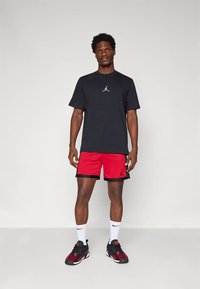 Mann steht vor einfarbigem Hintergrund und trägt ein schwarzes Jordan-T-Shirt, rot-schwarze Shorts, weiße Nike-Socken und schwarz-rote Turnschuhe.