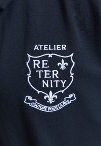 Tissu noir avec un emblème de bouclier brodé en blanc portant l'inscription "ATELIER RETERNITY" et une bannière en dessous indiquant "COUTURE POUR LA RUE" avec des symboles fleur-de-lis.