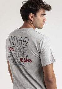 Camiseta de algodón gris con mangas cortas, que presenta texto en negrita en la parte posterior en azul y marrón que dice "1962 LOIS UNA DE LAS MARCAS DE VAQUEROS LÍDERES EN EL MUNDO."
