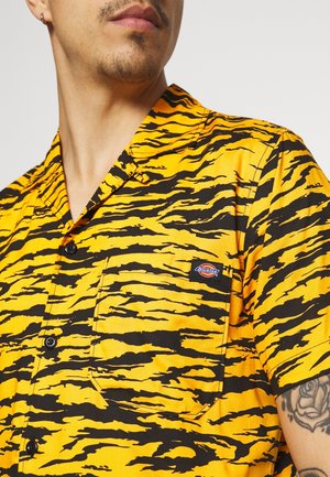 Mand iført en gul og sort tigerstribet skjorte med korte ærmer og en synlig tatovering på armen.