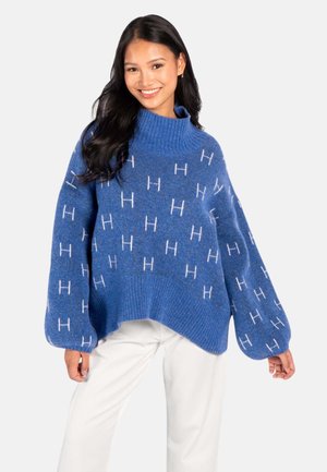 Ung kvinne med langt mørkt hår iført en blå, stor genser med hvite "H"-mønstre og hvite bukser, smilende mot kameraet.
