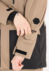 Beige ytterjacka med en svart sektion, med en kardborrestängning vid ärmarna och en hexagonal logotyp på ärmen. Slät, vattentålig tyg.