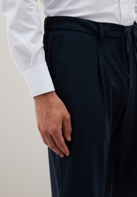 Männliche Hand, die neben einer marineblauen Hose mit Zugband ruht, getragen mit einem weißen Langarmhemd mit geknöpften Manschetten.