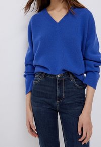 Pull bleu à col en V en tissu doux, porté par-dessus un jean en denim foncé à coupe ajustée. Le pull est pourvu de poignets et d'un ourlet côtelés.