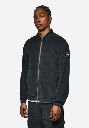 Schwarze Cordjacke mit einem Kragen, Frontreißverschluss, zwei Fronttaschen und elastischen Bündchen. Mit einem Logopatch am Ärmel.