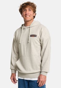 Mladý muž sa usmieva, má na sebe béžovú mikinu Billabong s predným vreckom a modré džínsy, stojí na jednoliatom pozadí.