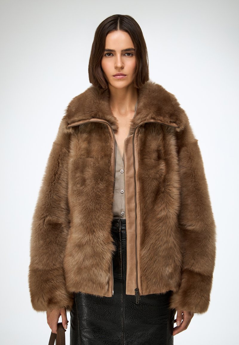 Bruine bontjas met een zachte, gestructureerde buitenkant, ritssluiting aan de voorkant en brede kraag, gecombineerd met een zwarte leren rok.