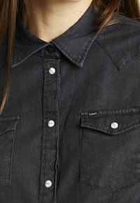 Tmavě modrá džínová košile s knoflíkovým zapínáním, špičatým límcem a dvěma náprsními kapsami. Obsahuje bílé akcenty knoflíků a malé logo na kapse.