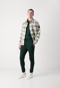 Rutete flanellskjorte i grå og krem, grønn genser, matchende joggebukser og hvite joggesko. Justerbar midje, frontlommer og tettsittende mansjetter.