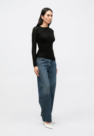 Bluză neagră cu mâneci lungi, textură plisată, combinată cu blugi evazați, cu talie înaltă, albastru deschis, uzat, și pantofi albi, cu vârf ascuțit.