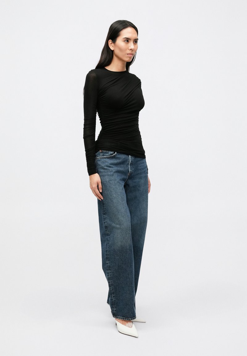 Sort langærmet top med rynket tekstur kombineret med højttaljede, falmede blå bukser med vide ben og hvide spidse hæle.