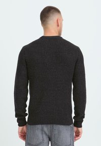 Mand med kort hår iført en sort struktureret striktrøje og grå jeans, vendt væk mod en ensfarvet lys baggrund.