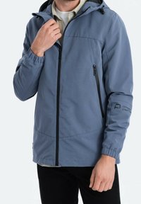 Homme portant une veste à capuche bleue zippée avec poches zippées et poignets élastiques, ajustant le col avec la main.