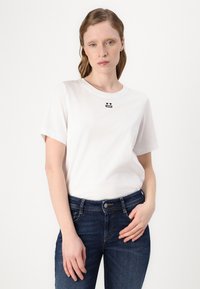 Weißes Baumwoll-T-Shirt mit kurzen Ärmeln, ausgestattet mit einem kleinen schwarzen grafischen Design auf der Brust. Kombiniert mit dunkelblauen, enganliegenden Jeans.