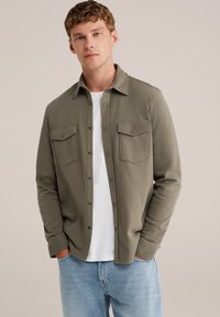 Cămașă verde măsliniu, cu butoniere, din material moale, cu două buzunare la piept și guler standard, purtată peste un tricou alb.