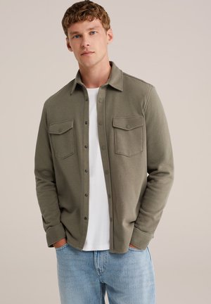 Camisa de botón verde oliva hecha de tela suave, con dos bolsillos en el pecho y un cuello estándar, usada sobre una camiseta blanca.
