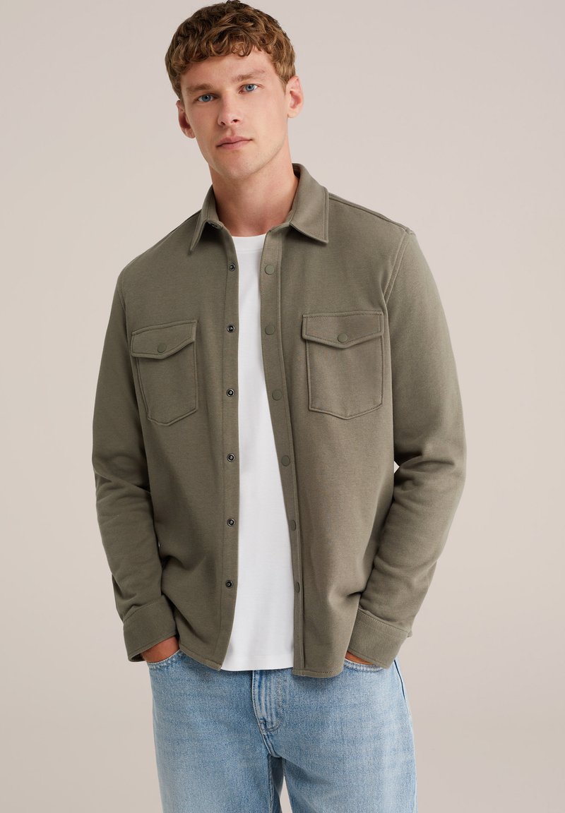 Cămașă verde măsliniu, cu butoniere, din material moale, cu două buzunare la piept și guler standard, purtată peste un tricou alb.