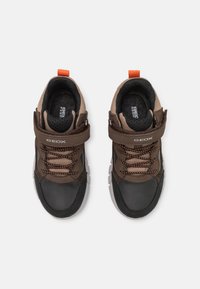 Zapatillas marrones con detalles en negro, material texturizado, tirador naranja, correa ajustable y cordones de contraste. Diseño ligero y transpirable.