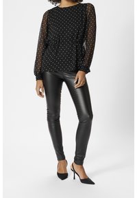 Blouse noire à pois avec manches longues transparentes, taille cintrée. Associée à des leggings en cuir noir ajustés et des escarpins noirs à bouts pointus.