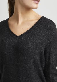 Pull en tricot noir, texturé, avec un décolleté en V, présentant un subtil scintillement et une coupe décontractée, mettant en avant une apparence douce et confortable.