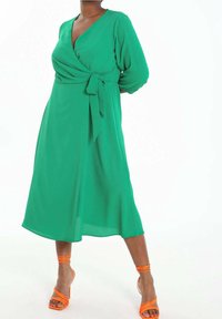 Grünes Wickelkleid mit drei Viertelärmeln, Taillierungsdetail, fließendem Stoff und Midi-Länge. Kombiniert mit orangefarbenen, schnürbaren Sandalen.