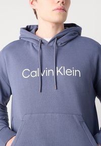 Felpa cappucciata in morbido tessuto blu testurizzato, con tasca a marsupio e logo "Calvin Klein" bianco sul davanti, arricchita da lacci con puntali in argento.
